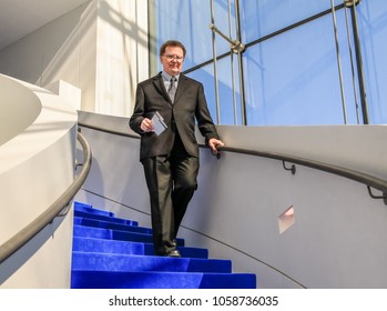 Smiling Older Man Dressed In A Suit Walking  Down The Staircase Of A Building With Large Glass Window; Blue Sky In The Background