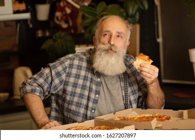 Smiling Older Man Eating Pizza Slice Sitting At Living Room. Bearded Senior Man Eating Italian Food.