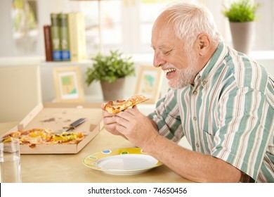 Smiling Older Man Eating Pizza Slice Sitting At Living Room Table.?