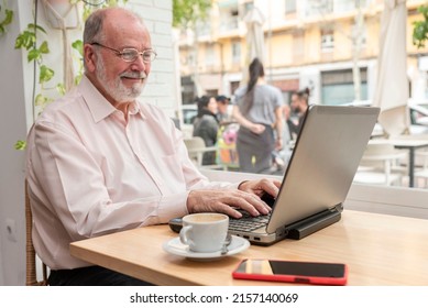 A Smiling Older Man With Eyeglasses Sitting In The Coffee Shop Bar Drinking Coffee And Working On His Laptop