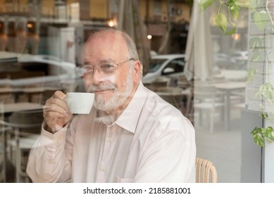 a smiling older man with glasses sitting behind a window in a coffee bar holding a cup of coffee - Powered by Shutterstock