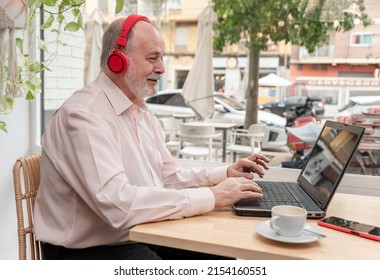 A Smiling Older Man With Headphones Looking At A Laptop Screen Watching A Seminar, Webinar Or Having Online Classes Sitting In A Coffee Shop, His Cup Of Coffee Next To Him