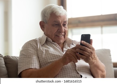 Smiling Older Man Holding Phone, Using Mobile Device Apps, Looking At Screen, Happy Mature Male Chatting Online, Texting, Writing Message, Having Fun With Gadget, Playing, Sitting On Couch