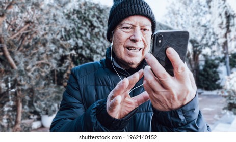 Smiling Older Man Makes A Video Call With His Black Smart Phone And White Headphones In His Backyard With A Background Of Plants