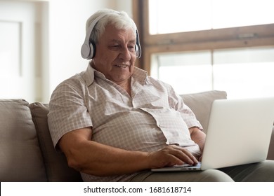 Smiling Older Man Wearing Headphones Using Laptop At Home, Looking At Screen, Listening To Favorite Music And Searching Information In Internet, Happy Mature Male Making Video Call, Sitting On Couch