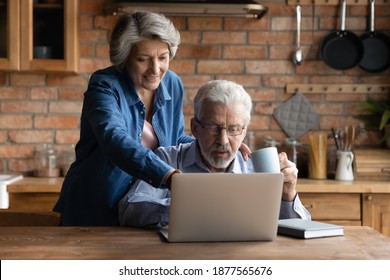 Smiling Older Mature Hoary Woman Pointing Finer At Laptop Screen, Choosing Holidays Trip With Relaxed Senior Husband Online. Happy Middle Aged Family Couple Using Modern Tech Gadget Together At Home.