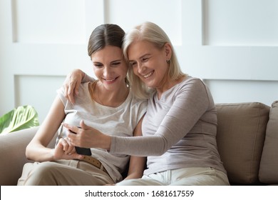 Smiling older mother and adult daughter using phone together, sitting on cozy sofa at home, happy young woman and mature mum looking at smartphone screen, watching video, having fun, two generations