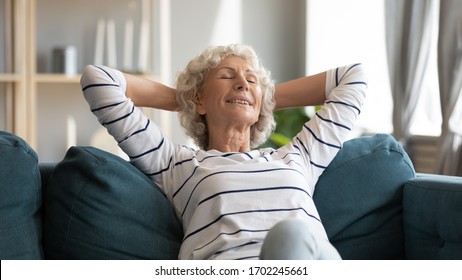 Smiling Older Woman Leaning Back, Relaxing On Cozy Couch, Happy Mature Senior Female With Hands Behind Head Sitting On Sofa In Living Room, Daydreaming With Closed Eyes, Lazy Peaceful Weekend