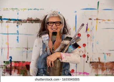 smiling older woman, proud artist, in her fifties with grey hair and black glasses and many paintbrushes