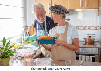 Smiling Older Woman Showing Her Freshly Baked Homemade Plumcake While Her Husband Washes The Dishes. Concept Of Family And Domestic Partnership