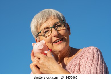 Smiling Older Woman With White Hair, Glasses, Pink Sweater And White Teeth With A Piggy Bank In Hands On Isolated Blue Background