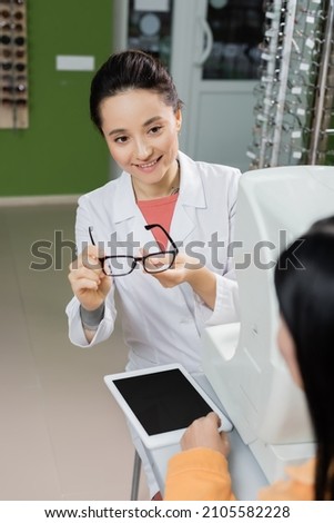 smiling ophthalmologist holding eyeglasses near blurred woman, digital tablet and vision screener in optics shop