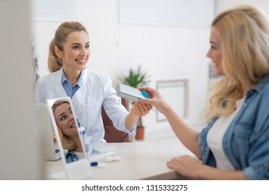 Smiling Optician Sitting At The Table And Giving Silver Packaging Box With Contact Lenses To Woman