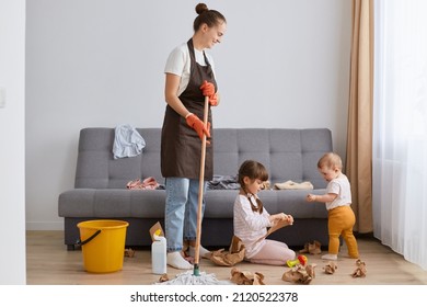 Smiling optimistic woman with bun hairstyle wearing white t shirt, brown apron and jeans, cleaning house, posing with her little children, young housewife washing floor and looking after kids.