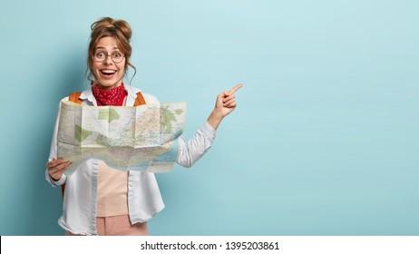 Smiling Optimistic Young European Female Guide Holds Map, Shows Direction To Tourist Away, Demonstrates Destinations Of Showplace, Dressed Casually, Isolated On Blue Wall, Searches Right Route