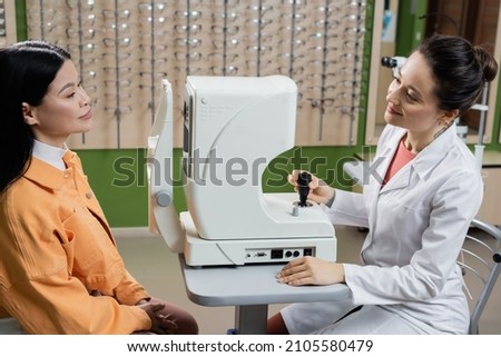smiling optometrist looking at asian woman near vision screener and blurred assortment of eyeglasses