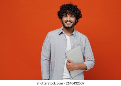 Smiling overjoyed excited happy jubilant young bearded Indian man 20s years old wears blue shirt hold use work on laptop pc computer looking camera isolated on plain orange background studio portrait