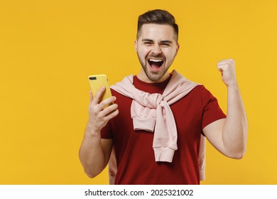 Smiling Overjoyed Young Man Wear Red T-shirt Casual Clothes Use Mobile Cell Phone Do Winner Gesture Clench Fist Isolated On Plain Yellow Color Wall Background Studio Portrait. People Lifestyle Concept