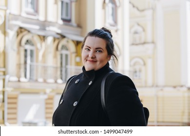 Smiling overweight brunette woman in a stylish black coat. Tourist student walking around with a backpack. Portrait of happy smiling brunette plus size girl.
