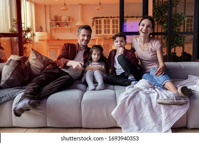 Smiling Parent With Kid Watching Movie On Tv In Cozy Home Living Room. Father, Mother And Children With Snack Sitting On Comfortable Sofa Front Of Tv-set. Happy Family Enjoy Weekend Together