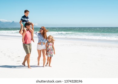 Smiling parents with children at sea. Cute son sitting on father shoulder with mother and sister walking while talking. Happy family with two children enjoying summer holiday at beach with copy space.