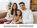 Smiling parents embracing their daughter while sitting together on a comfortable sofa in their modern living room, enjoying quality time as a family