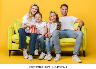 Smiling Parents And Kids Watching Movie On Sofa With Popcorn Bucket On Yellow