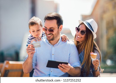 Smiling Parents And Little Girl With Tablet Pc And Credit Card Outdoor