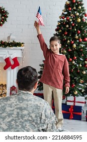 Smiling Patriotic Boy Standing With Usa Flag Near Christmas Tree And Dad In Camouflage On Blurred Foreground