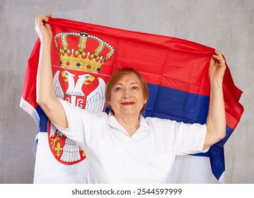 Smiling patriotic senior woman waving national flag of Serbia posing against gray studio background - Powered by Shutterstock