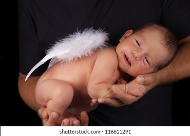 Smiling Peaceful Newborn Baby Boy With Angel Wings In Father's Hands. Studio Shot, Over Black Background.