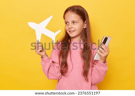 Smiling pensive little girl standing with smartphone and paper plane in hands booking tickets for raveling abroad choosing most comfortable airline posing isolated over yellow background.