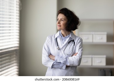 Smiling Pensive Older Female Doctor Standing By Window In Office, Looking Away, Thinking Of Patients, Medical Career Growth, Success. Practitioner With Hands Folded Head Shot Portrait