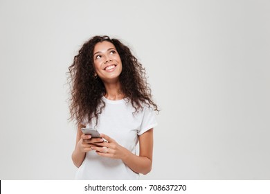 Smiling pensive woman holding smartphone in hands and looking up over gray background
