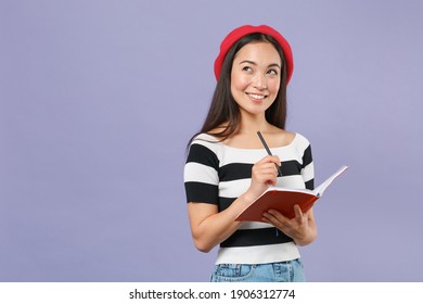 Smiling Pensive Young Brunette Asian Student Woman 20s Wearing Striped T-shirt Red Beret Standing Writing Notes In Notebook Looking Up Isolated On Pastel Violet Colour Background Studio Portrait