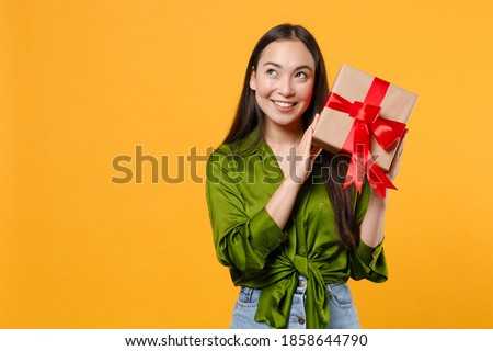Smiling pensive young brunette asian woman wearing basic green shirt standing hold red present box with gift ribbon bow looking aside up isolated on bright yellow colour background, studio portrait