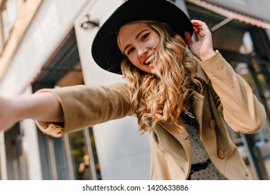 Smiling, perky curly young lady takes a selfie on the street. Portrait of a blonde with a gentle make-up in a hat - Shutterstock ID 1420633886