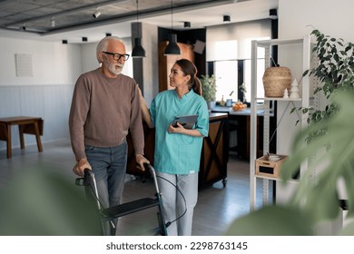 Smiling physiotherapist showing empathy and support to elderly male patient with disability while he's walking with walking frame. Physiotherapist supporting senior man. Physiotherapist in home visit. - Powered by Shutterstock