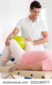 Smiling Physiotherapist Supporting Patient Squeezing Yellow Ball Between Knees