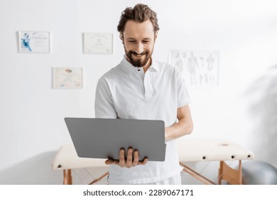 smiling physiotherapist using laptop in consulting room of rehabilitation center - Powered by Shutterstock