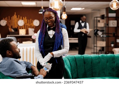 Smiling pleasant African American woman waitress holding POS terminal receiving cashless payment for food and drinks from hotel guest waiting for check-in in hotel lobby. NFC Technology in hospitality