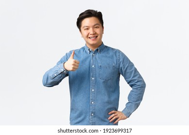Smiling Pleased Asian Male Student With Braces, Showing Thumbs-up, Recommend Product Or Service With Excellent Quality, Like And Approve Idea. Man Nod In Approval, Agree With Person, White Background
