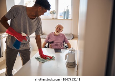 Smiling pleased disabled aged Caucasian person looking at the volunteer dusting the table in his presence