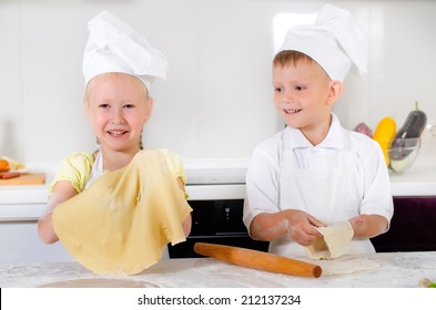 Smiling Pleased Little Girl Dressed In An Apron And Chefs Cap Holding Up Her Thinly Rolled Out Pastry For A Pizza Base With A Proud Smile Watched By Her Young Brother