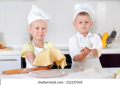 Smiling Pleased Little Girl Dressed In An Apron And Chefs Cap Holding Up Her Thinly Rolled Out Pastry For A Pizza Base With A Proud Smile Watched By Her Young Brother