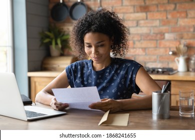 Smiling pleased young african american woman reading paper letter with good news, sitting at table with computer at home. Happy curious pretty millennial mixed race lady getting correspondence.