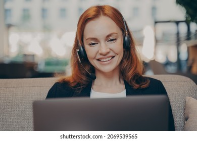 Smiling pleased young redhead woman wearing headset talking with colleagues online on laptop while sitting on sofa in modern office, participating in online conference during remote work from home - Powered by Shutterstock