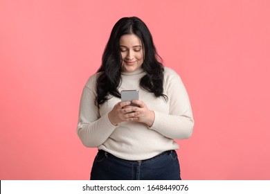 Smiling plump woman holding smartphone in hands, texting, reading message, or using new application, isolated on pink background with free space