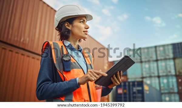 Smiling Portrait of a Beautiful Latin Female\
Industrial Engineer in White Hard Hat, High-Visibility Vest Working\
on Tablet Computer. Inspector or Safety Supervisor in Container\
Terminal.