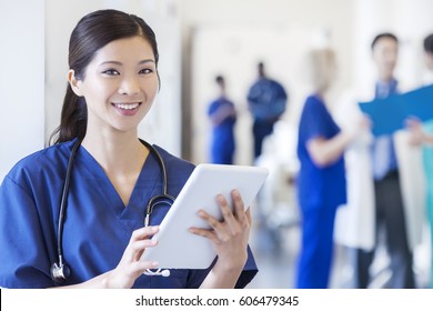 Smiling portrait ethnic female nursing staff wearing uniform scrubs using tablet technology in hospital corridor with clinical team